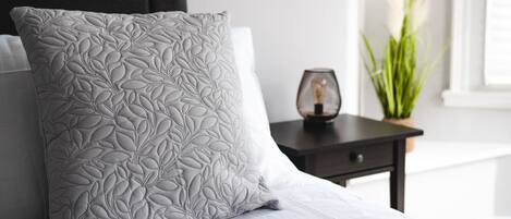 A neatly made bed with a patterned pillow and a bedside table with a lamp and a plant.