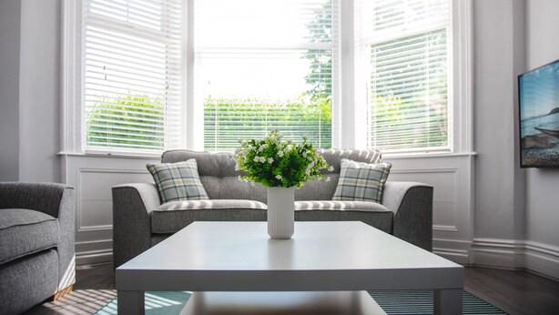 A room with a white table, grey sofa, and a window with white blinds. - Oxford House (Llandudno)