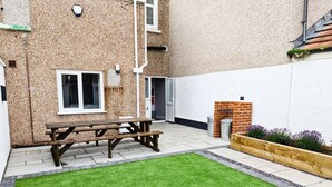 A patio area with a wooden picnic table, a small brick fireplace, and a well-maintained lawn. - Oxford House (Llandudno)