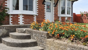 A brick house with white-framed windows, a stone staircase, and a flower bed with orange flowers. - Silver Crooks (Holsworthy)