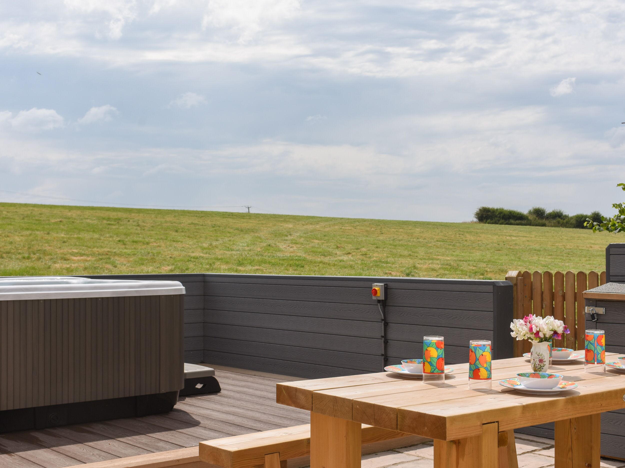 An outdoor patio with a wooden table set for dining, a hot tub, and a view of a grassy field.