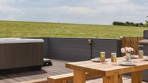 An outdoor patio with a wooden table set for dining, a hot tub, and a view of a grassy field. - Silver Crooks (Holsworthy)