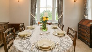 A dining room with a round table set for four, surrounded by wooden chairs. There is a vase with yellow flowers on the table, and the room has a window with curtains. - Silver Crooks (Holsworthy)