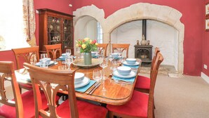 A dining room with a wooden table set for six, red upholstered chairs, a vase of flowers, and a stone fireplace with a wood-burning stove.