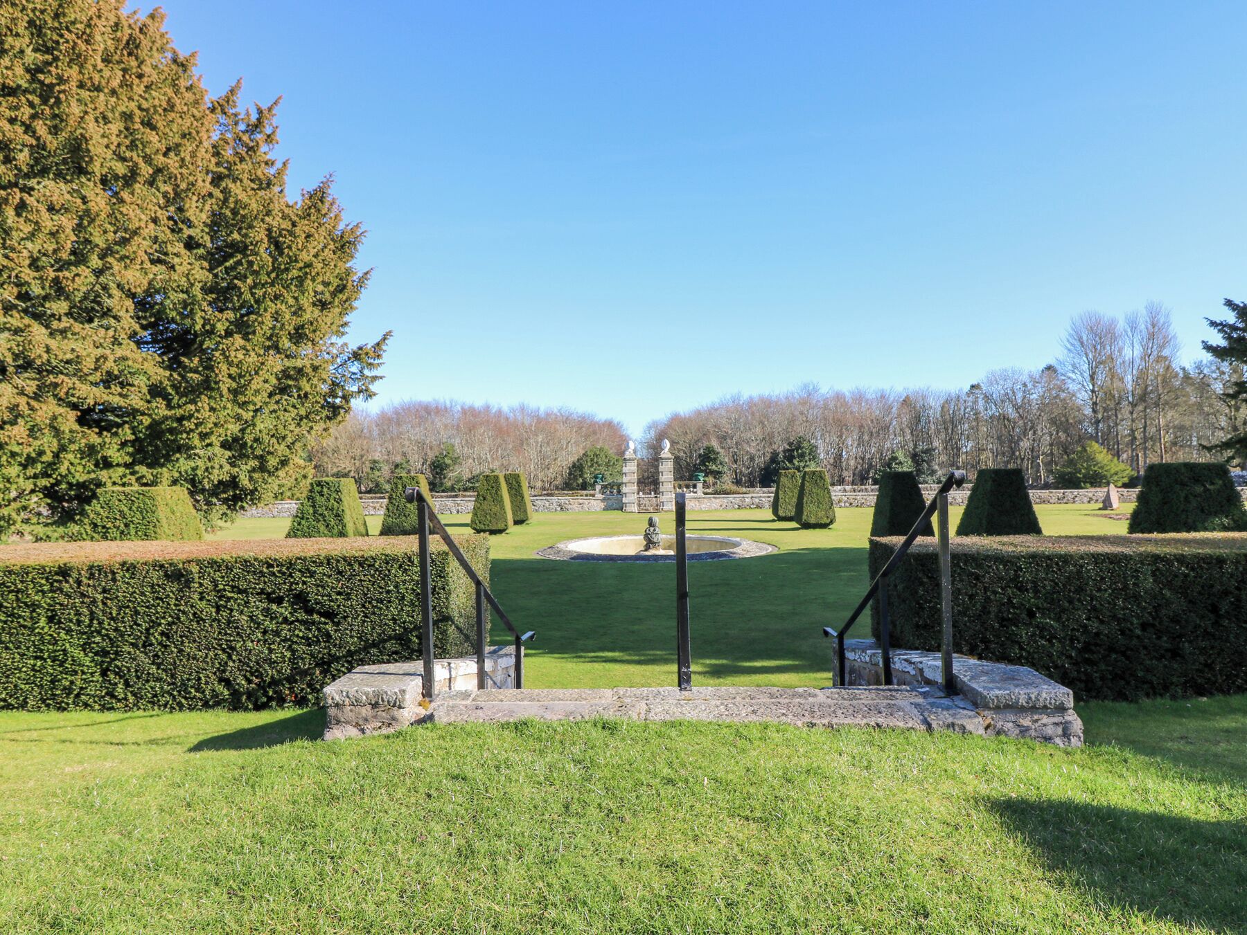 A well-maintained garden with neatly trimmed hedges, a central fountain, and a stone pathway leading through the greenery.