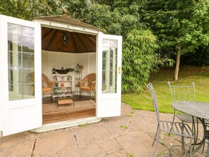 Una estructura pequeña y abierta con techo de madera y exterior blanco, que incluye mobiliario de mimbre, una mesa de vidrio y una silla de metal.