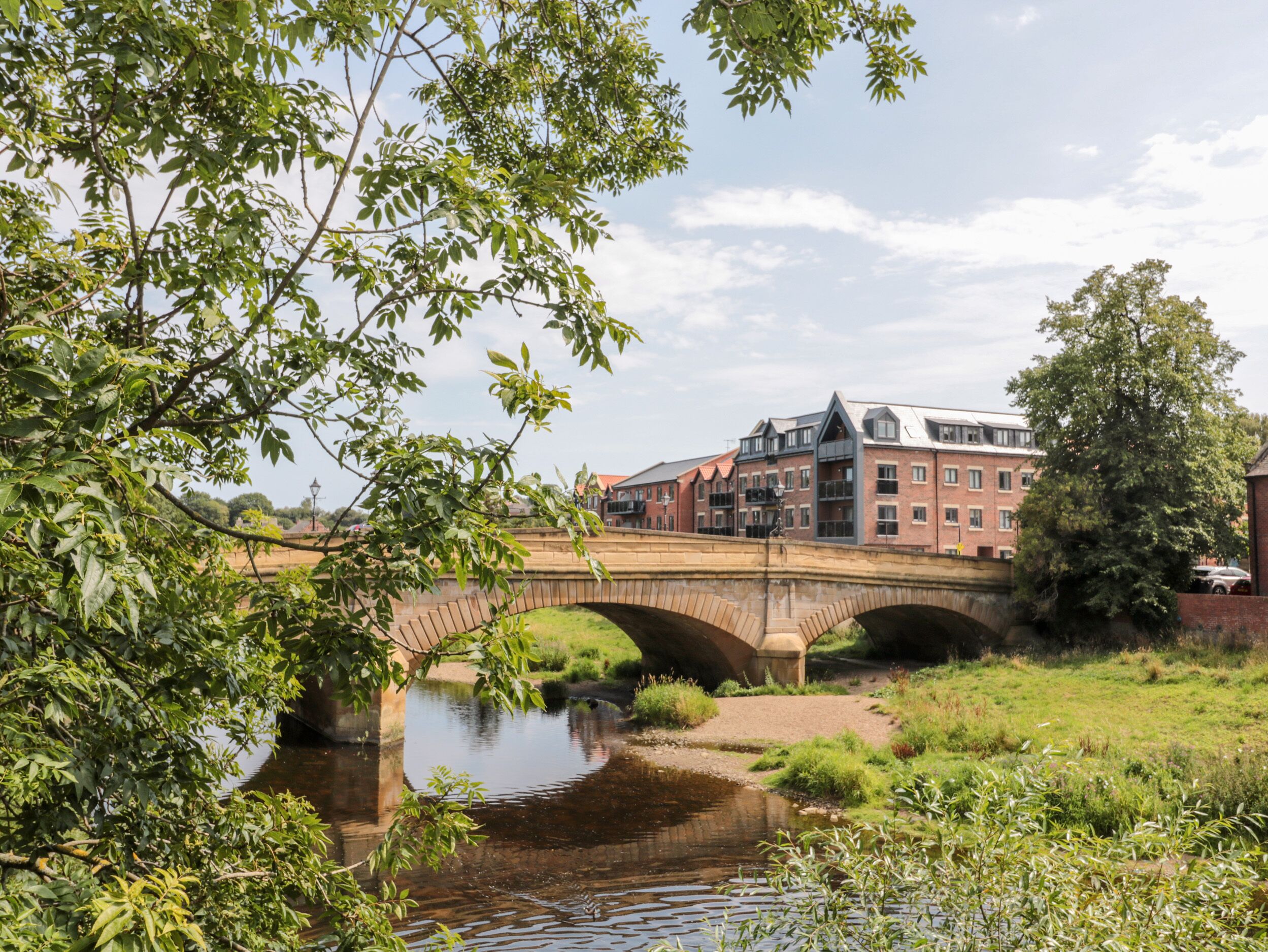 Eine historische Steinbrücke über einem ruhigen Fluss, mit üppiger Vegetation und einem traditionellen Backsteinhaus im Hintergrund.