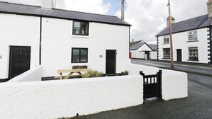 A white building with a black door, a window, and a small garden area with a bench and plants. - Menai Cottage (Llanfairpwllgwyngyll)