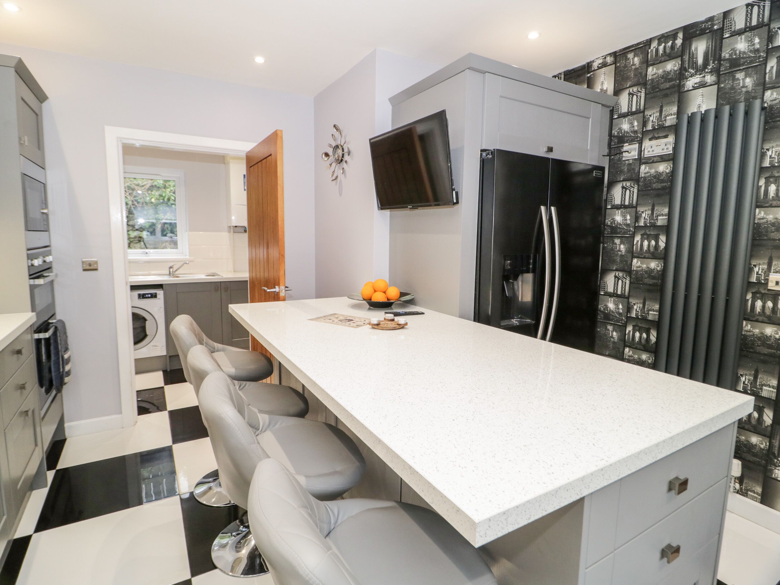 A modern kitchen with a white countertop, bar stools, a black refrigerator, and a wall-mounted TV.