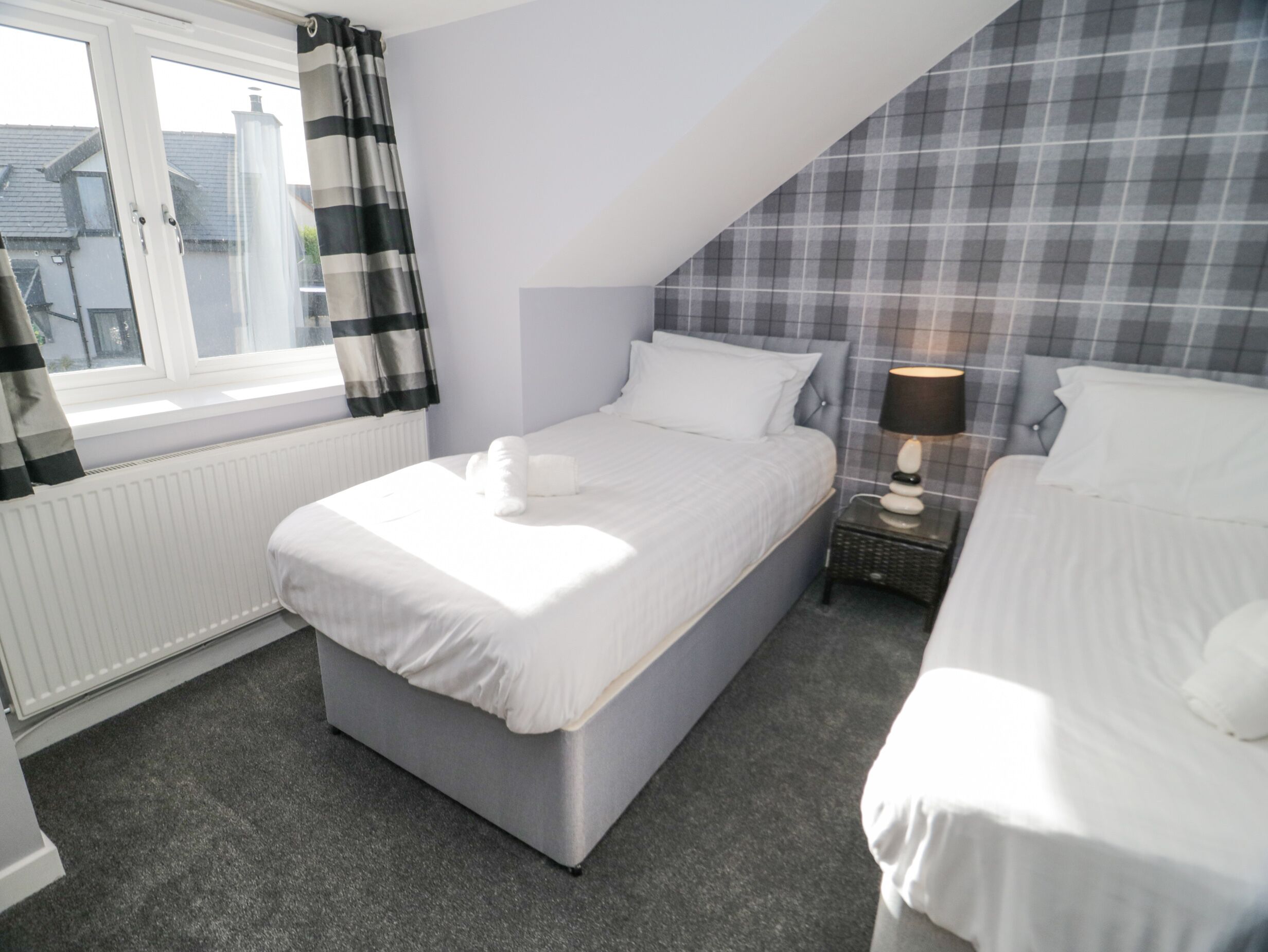 A bedroom with a bed, a bedside table with a lamp, a window with curtains, and a radiator.