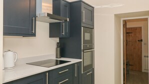 A modern kitchen with dark cabinets, a stainless steel range hood, a built-in oven, and a white countertop. - 3 Old Hall Cottages (Bakewell)