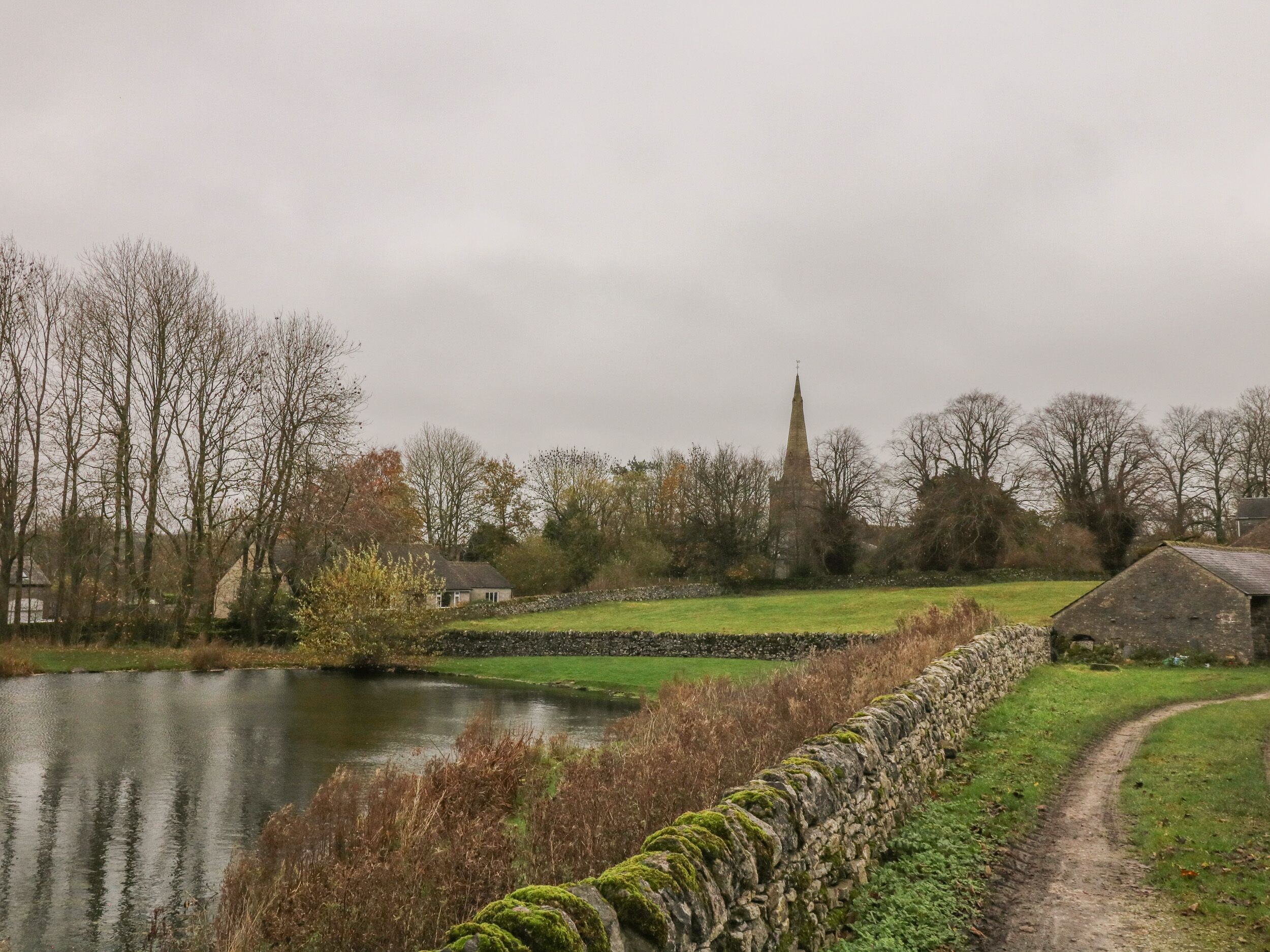 A rural landscape with a stone wall, a pond, and a church spire in the distance.