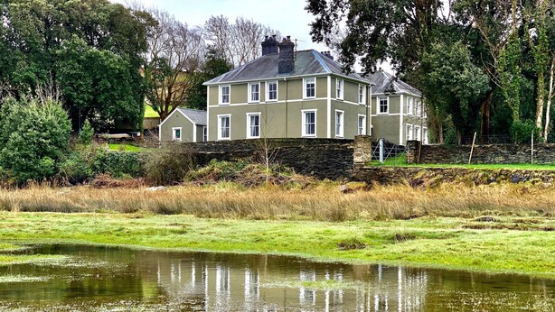 A large, two-story house with a stone foundation, surrounded by a grassy field and a pond. - Mor Edrin (Talsarnau)