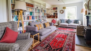 A cozy living room with a grey sofa, a red armchair, and a patterned rug. There are bookshelves filled with books and decorative items. A fireplace is visible in the background. - Mor Edrin (Talsarnau)