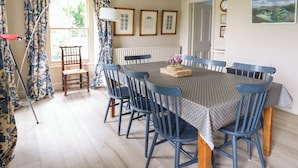 A dining room with a round table, blue chairs, floral curtains, and framed pictures on the wall. - Mor Edrin (Talsarnau)