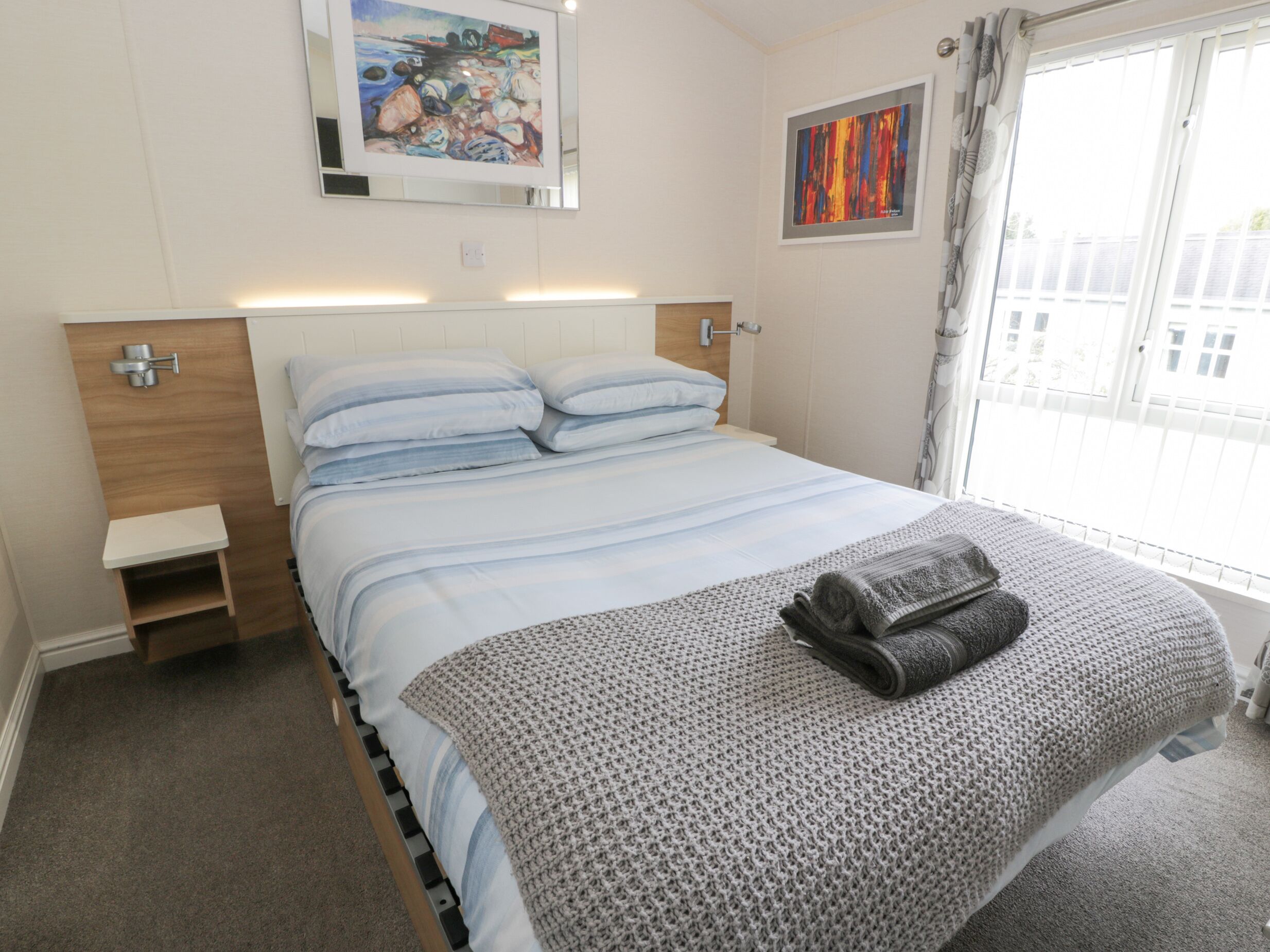 A neatly made bed with a grey knitted blanket and pillows, a small bedside table, and artwork on the walls.
