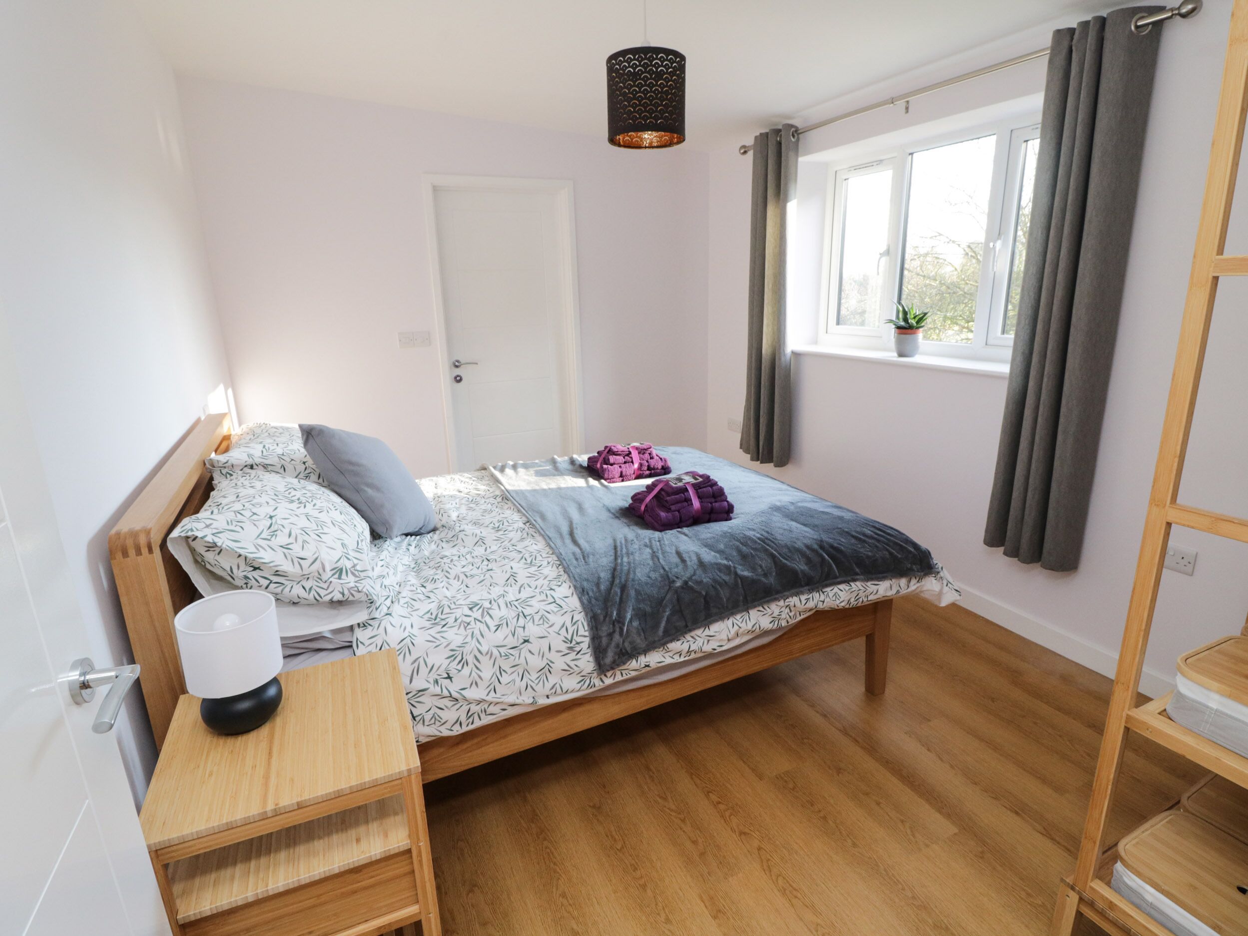 Een keurig opgemaakt slaapkamer met een houten bed, een nachtkastje, een raam met gordijnen en een kleine potplant op de vensterbank.