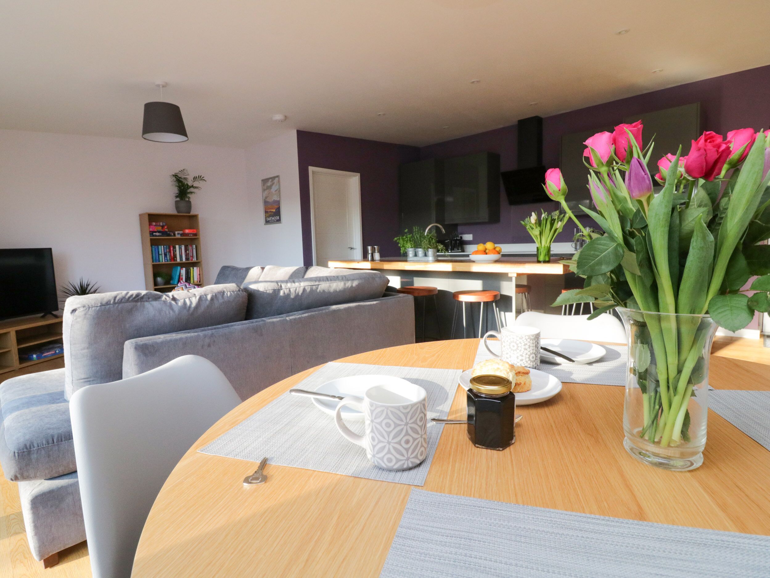 Una sala de estar moderna con un sofá gris, una mesa de comedor con un jarrón de flores y una cocina al fondo.