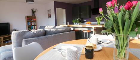 A modern living room with a grey sofa, a dining table with a vase of flowers, and a kitchen in the background.