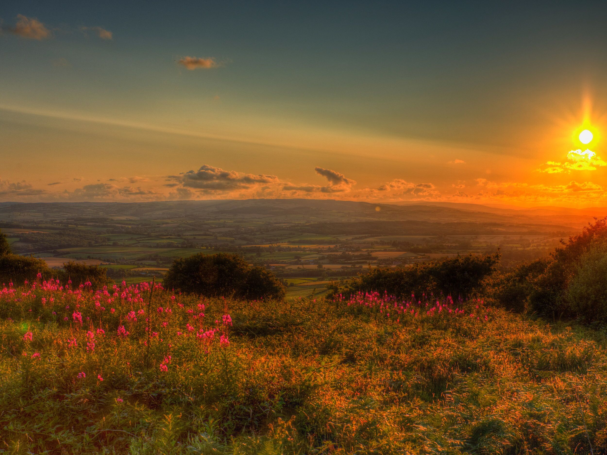 A landscape with a sunset, fields, and distant hills.