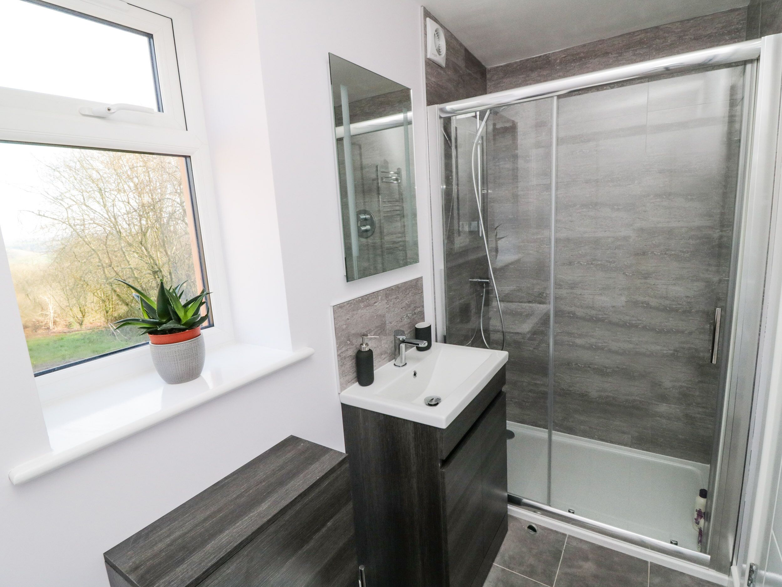 Een moderne badkamer met een douchecabine van glas, een wastafel met spiegel erboven en een potplant op een vensterbank.