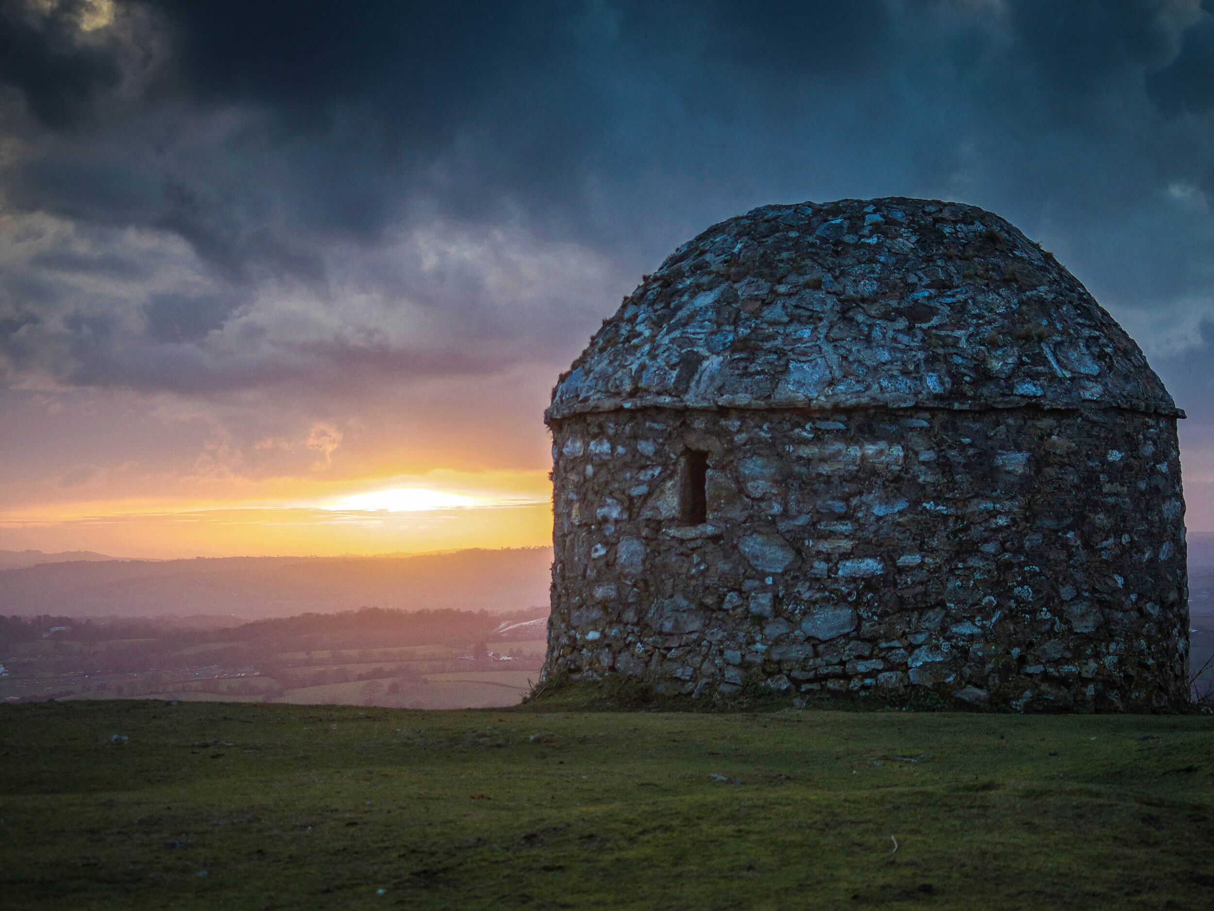 A stone building with a dome roof, surrounded by a grassy field and a hilly landscape in the background.
