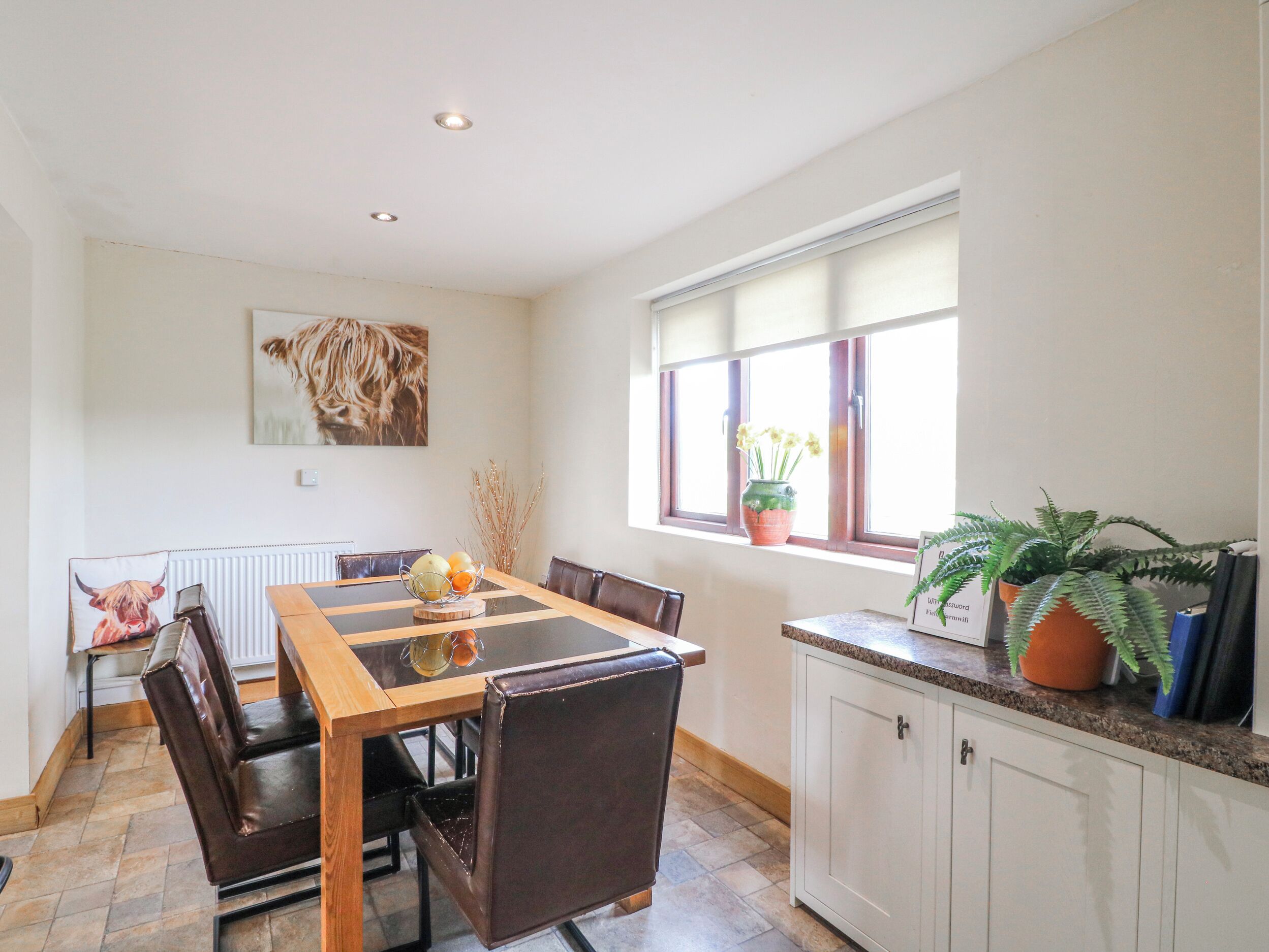 A dining area with a wooden table, chairs, a radiator, a window with plants, and a painting on the wall.
