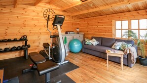 Interior de una cabaña de madera con una bicicleta estática, pesas, un sofá y una ventana.