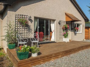 A house with a wooden deck, two white metal chairs, and potted plants with flowers. - Beechwood Cottage (Pitlochry)