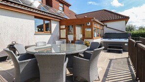 En terrasse med et rundt glasbord og fletstole, grill og et overdækket siddeområde med træbjælker.