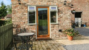 A brick building with a wooden door and white-framed windows. There is a small patio with a table and chairs, and potted plants. - Decca Cottage Nidd Grange (York)