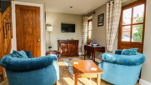 A living room with two blue armchairs, a wooden coffee table, a wooden cabinet, a television, and a window with curtains. - Decca Cottage Nidd Grange (York)