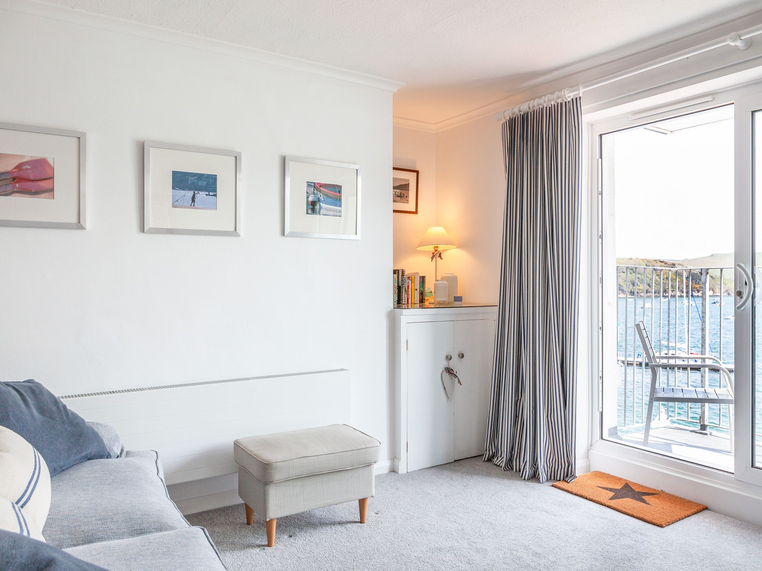 Un salon moderne avec un canapé gris, un petit pouf, un meuble blanc et une vue sur la mer, visible à travers une grande fenêtre ornée de rideaux à rayures.