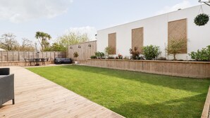 Un jardin avec une terrasse en bois, une pelouse verdoyante, un espace détente avec une table et des chaises, et un bâtiment blanc doté de fenêtres et d’une plante suspendue.