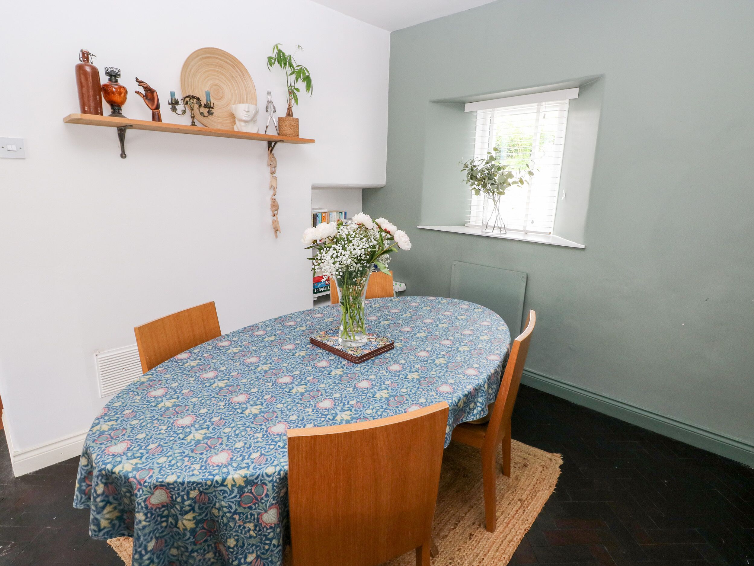 A dining area with a round table covered by a floral tablecloth, wooden chairs, a woven rug, and a window with a plant.