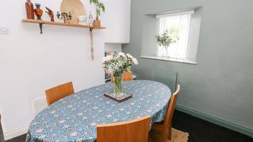 A dining area with a round table covered by a floral tablecloth, wooden chairs, a woven rug, and a window with a plant.