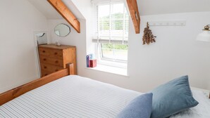 Een slaapkamer met een bed, een houten dressoir, een raam met zonwering, en een klein plankje met boeken.