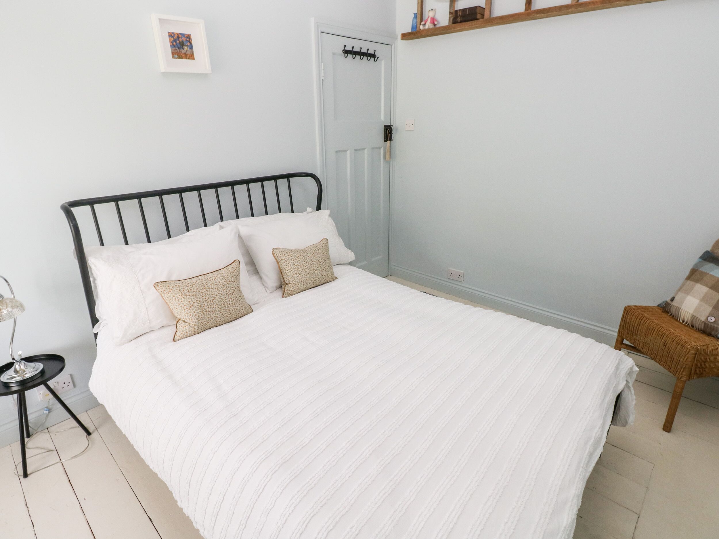 Een net opgemaakt bed met een witte dekbedovertrek en twee gekleurde kussens, een klein nachtkastje met een lamp, en een rieten stoel.