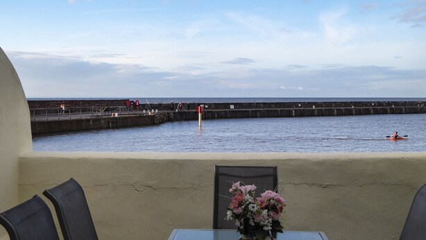 A table with two wine glasses and a bottle, a flower vase, and a view of the sea and a pier with people. - Harbour Cottage (Watchet)