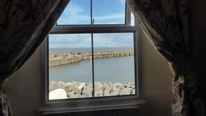A room with a large window overlooking a rocky shoreline and a calm sea. - Harbour Cottage (Watchet)