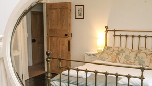 A bedroom with a wooden door, a bedside table with a lamp, a bed with a metal headboard, and a window with a view of the outside. - Harbour Cottage (Watchet)
