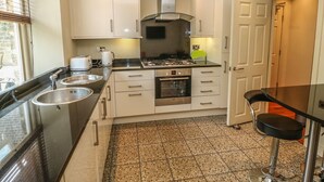 Cocina moderna con estufa a gas, horno y pileta doble. Hay un taburete de bar y una ventana con persianas.