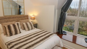 A bedroom with a large bed, a nightstand with a lamp, a mirror, and a window with curtains. - Yr Hen Ysgol, The Old School (Porthmadog)