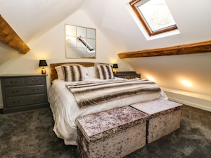 Quarto com cama, mesas de cabeceira, penteadeira, um banco, um claraboia e um espelho na parede.