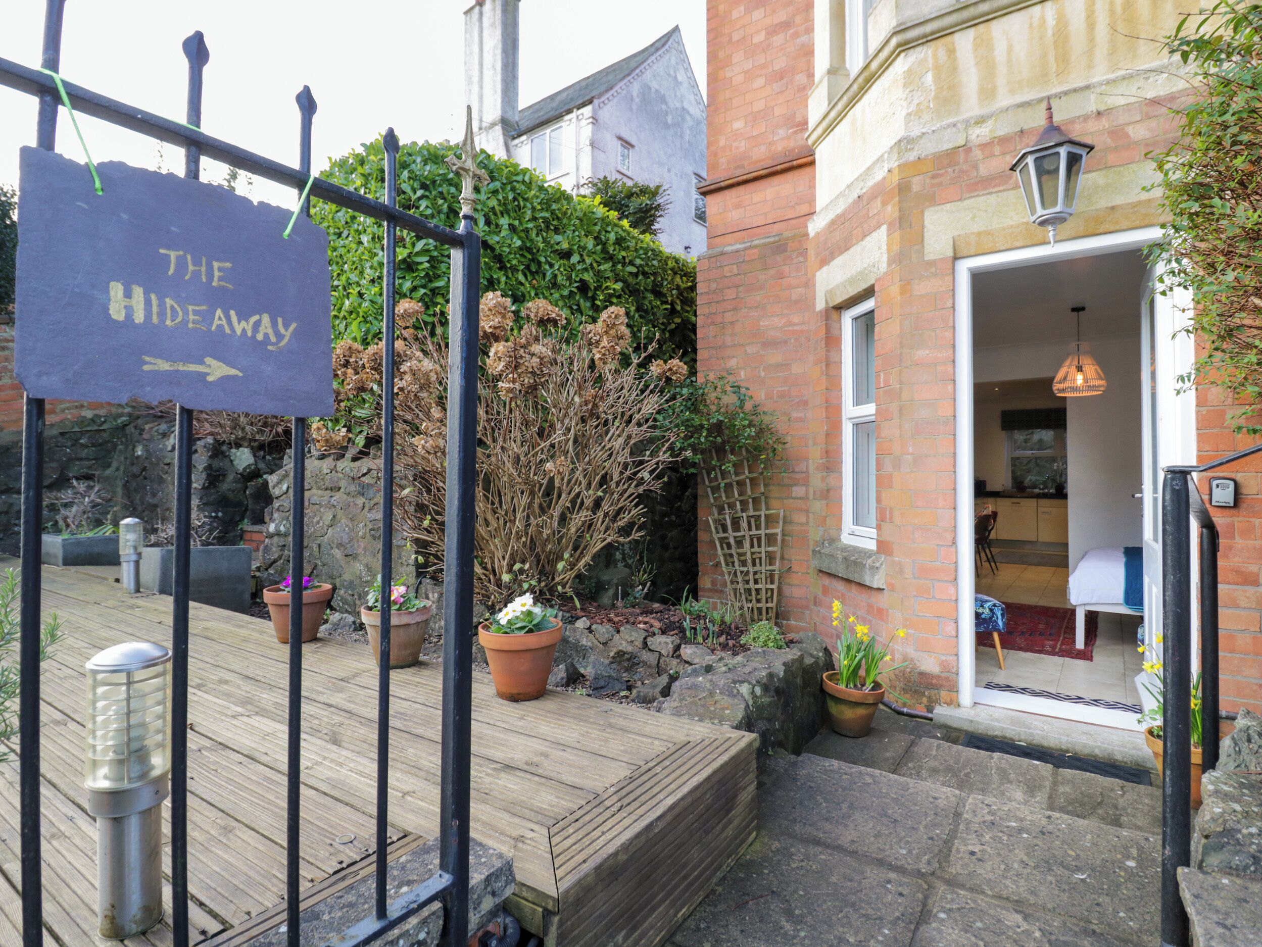 A wooden deck with a sign reading "The Hideaway," potted plants, a stone wall, and a brick building with a door and window.
