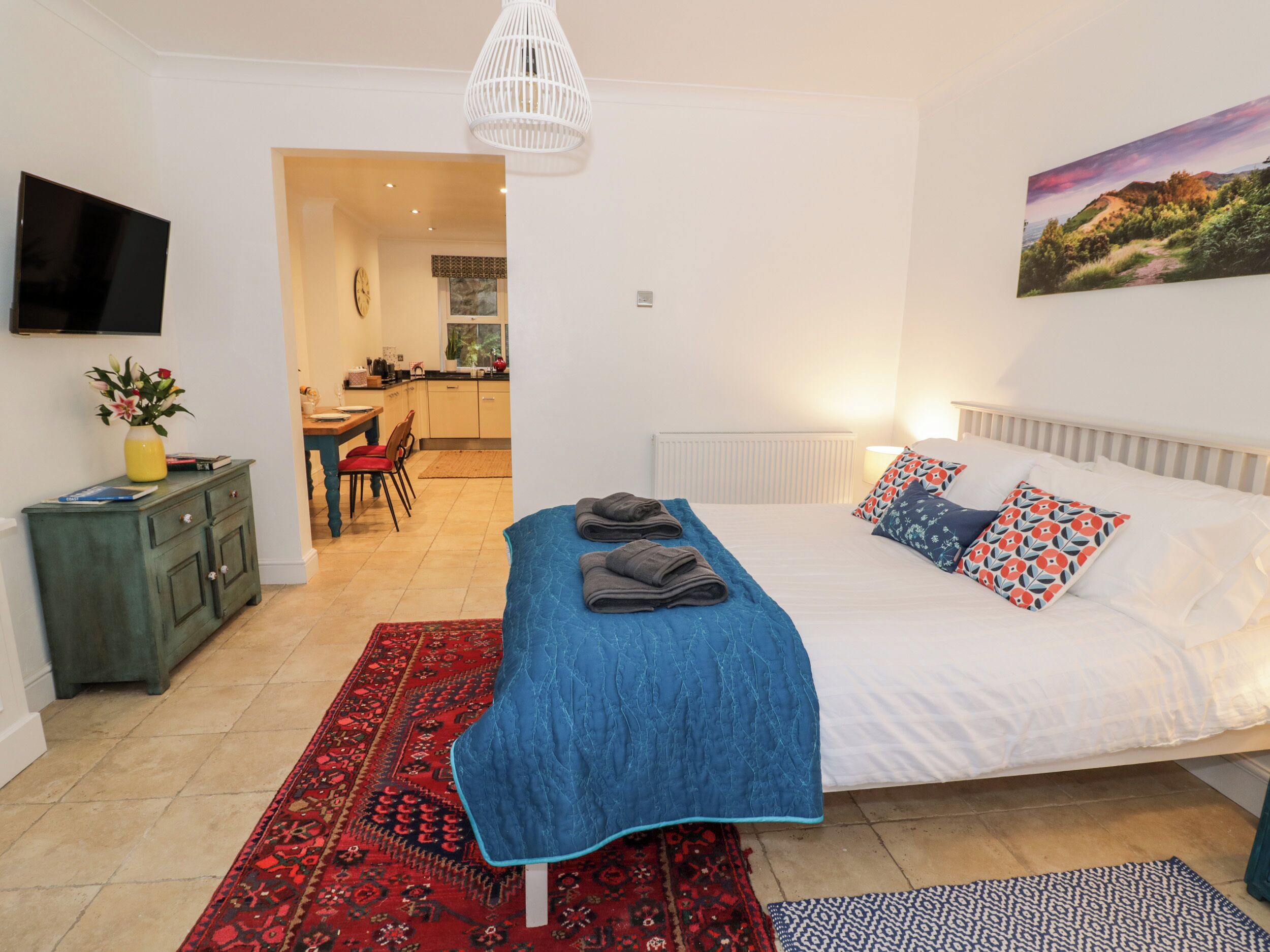 Un dormitorio con una cama, un tapete rojo y azul con estampado, una pared blanca y un cuadro de un paisaje.