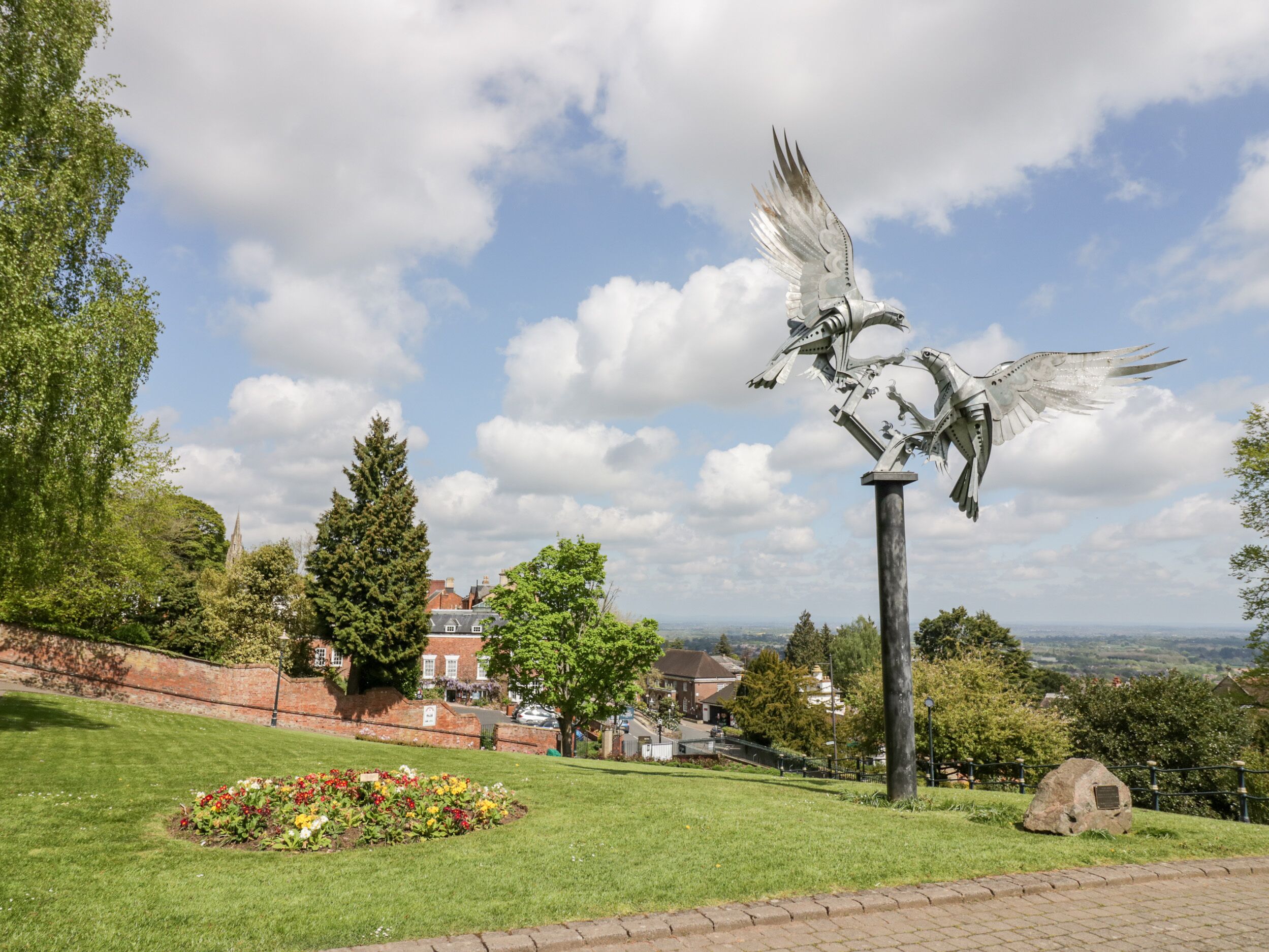 Una escultura de pájaros sobre un pedestal alto en un parque, con vistas a casas al fondo.