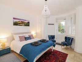 Quarto com cama, manta azul, almofadas estampadas, mesa de cabeceira com abajur, tapete vermelho, luminária branca no teto, janela com persianas e cadeira com padrão azul.