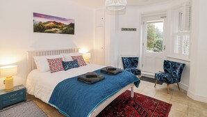 Une chambre à coucher avec un lit, une couverture bleue, des coussins à motifs, une table de chevet avec une lampe, un tapis rouge, un luminaire blanc au plafond, une fenêtre avec des volets et une chaise à motifs bleus.