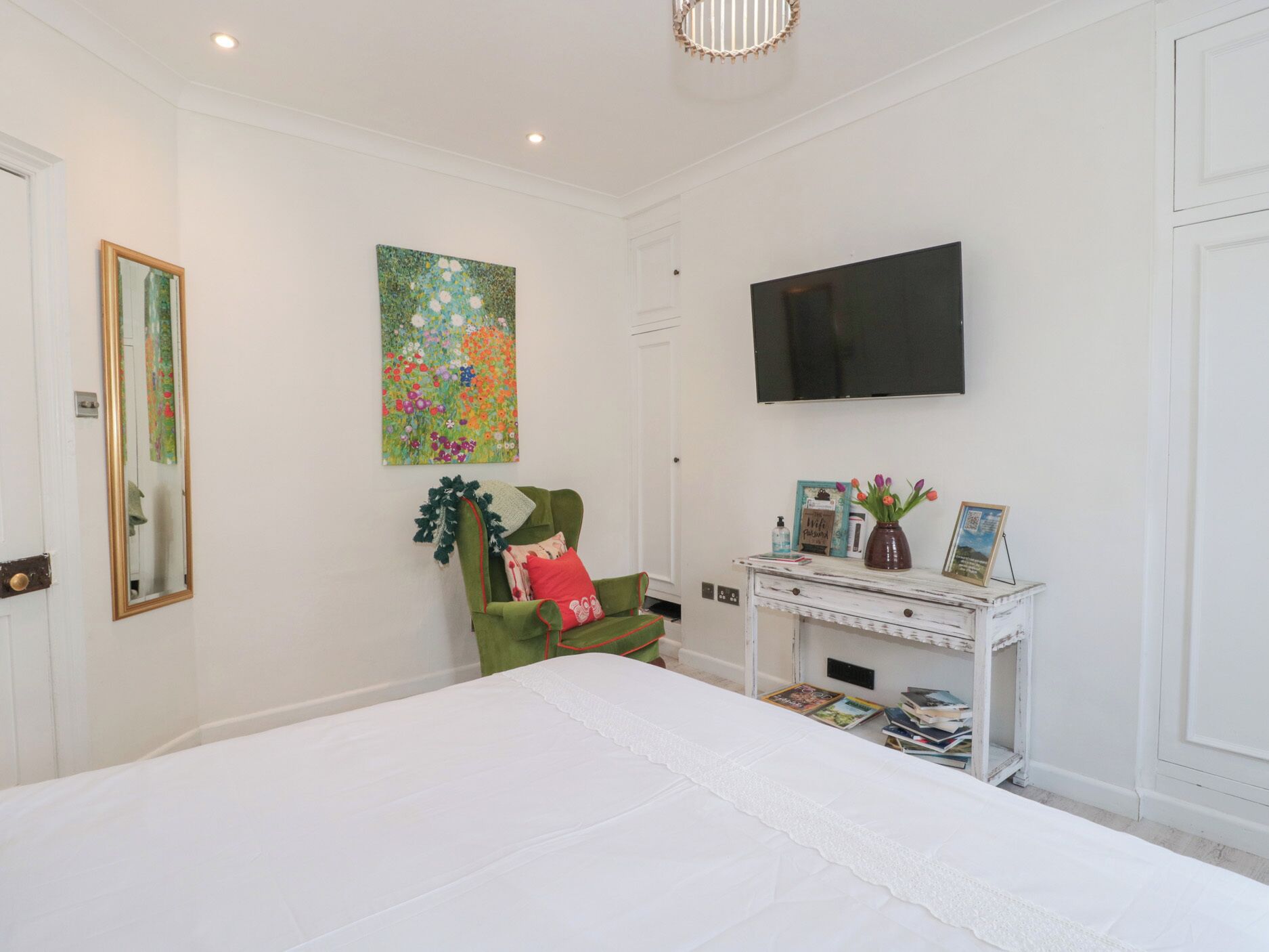 A bedroom with a large bed, a green armchair, a white dresser, a flat-screen TV, and a floral painting on the wall.